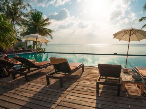 Poolside scene with lounge chairs, umbrellas, and palm trees by an infinity pool overlooking the ocean under a cloudy sky.