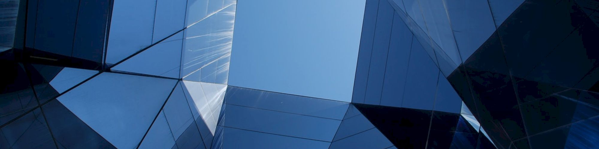 The image shows a view looking up at intersecting reflective glass structures against a blue sky, creating a geometric, abstract pattern.