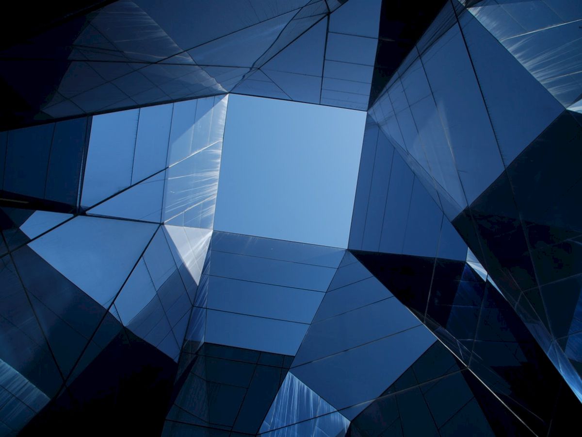 A view up through angular, reflective glass structures creating a geometric frame around a patch of clear blue sky.