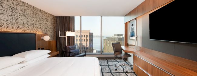 A modern hotel room with a large bed, wall-mounted TV, and a desk. Large windows provide a view of city buildings and water.