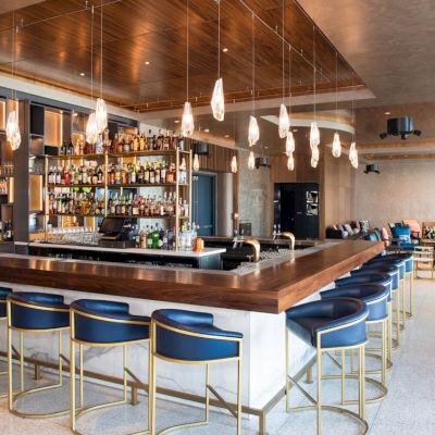 A modern bar with blue stools, pendant lighting, and a fully stocked back wall in a sleek lounge area.