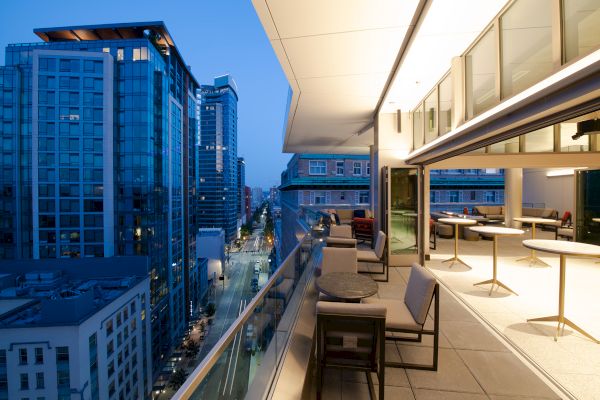 A modern balcony with seating overlooks a city street lined with tall buildings at dusk, featuring a sleek, open interior space.