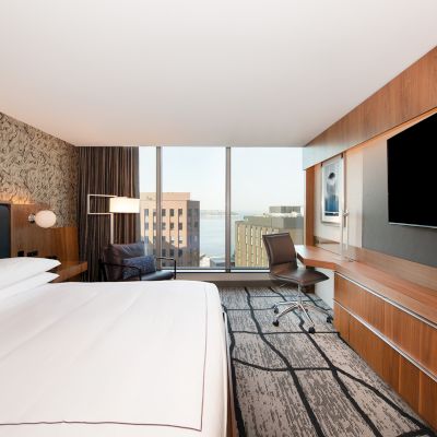 A modern hotel room with a large bed, desk, TV, and a window view of buildings and water.