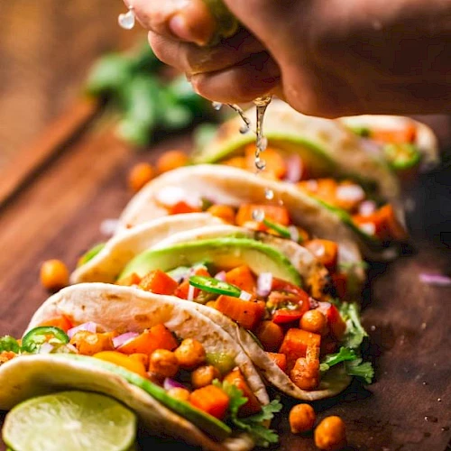 A hand squeezes lime juice over tacos filled with chickpeas, avocado, and veggies, garnished with cilantro, on a wooden board.
