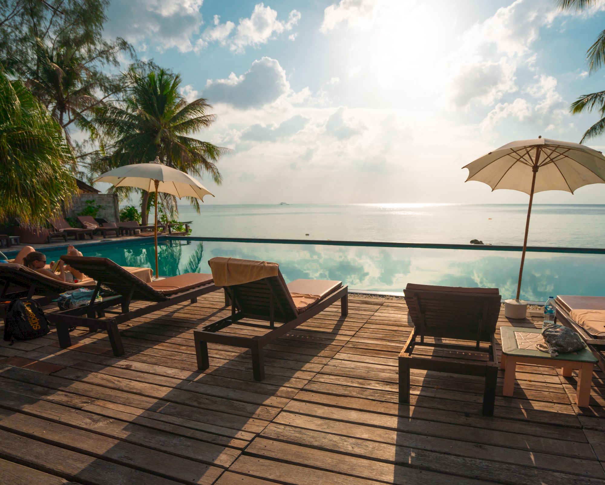 A serene tropical beach scene with sun loungers on a wooden deck, overlooking a calm sea and sky, with umbrellas and palm trees.