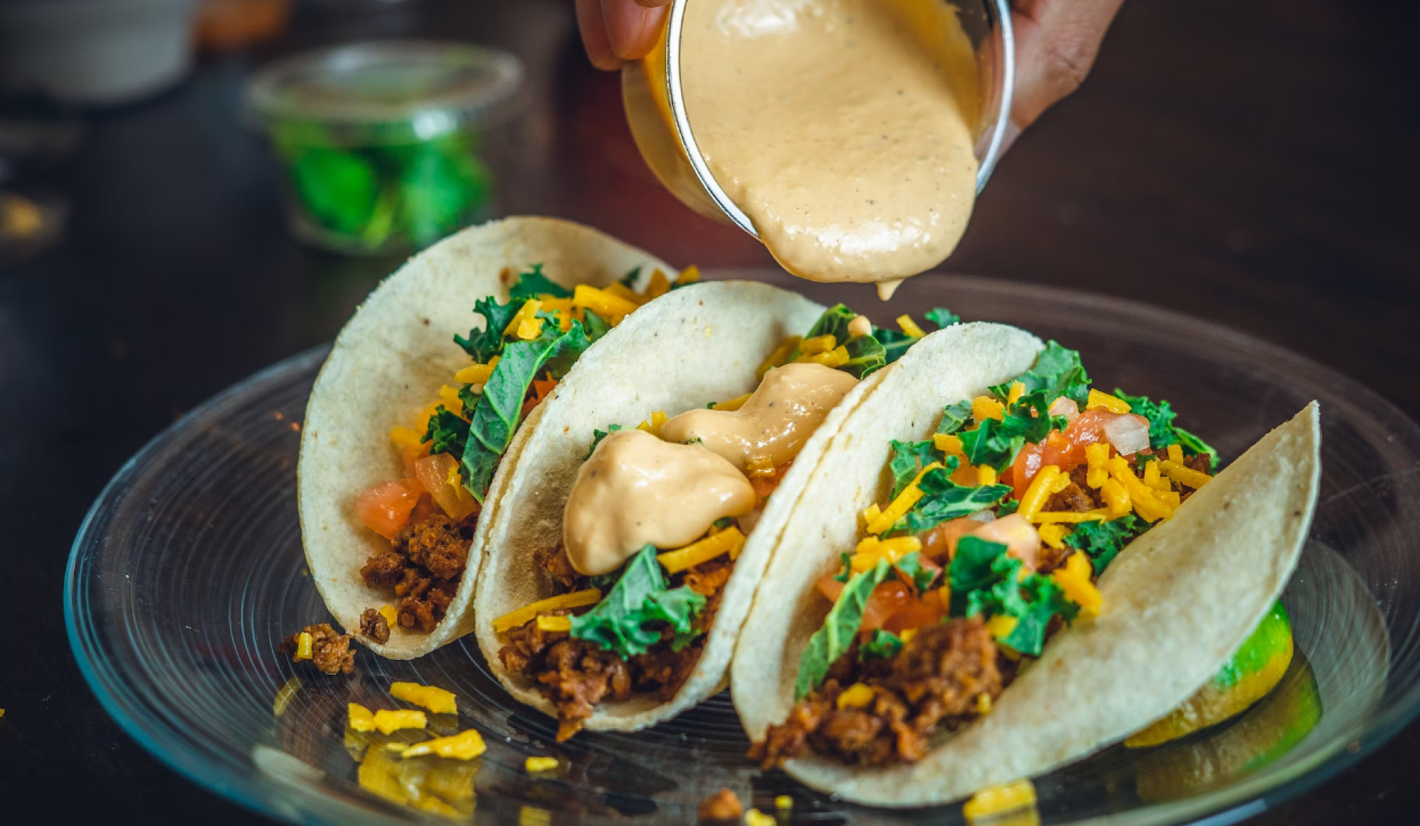 Three tacos with vegetables, cheese, and a creamy sauce being poured over them on a glass plate.