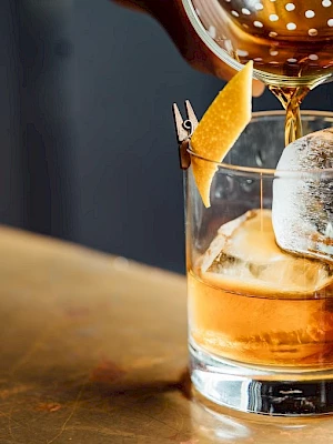A cocktail being poured into a glass with ice and a twist of lemon peel garnish on a table.