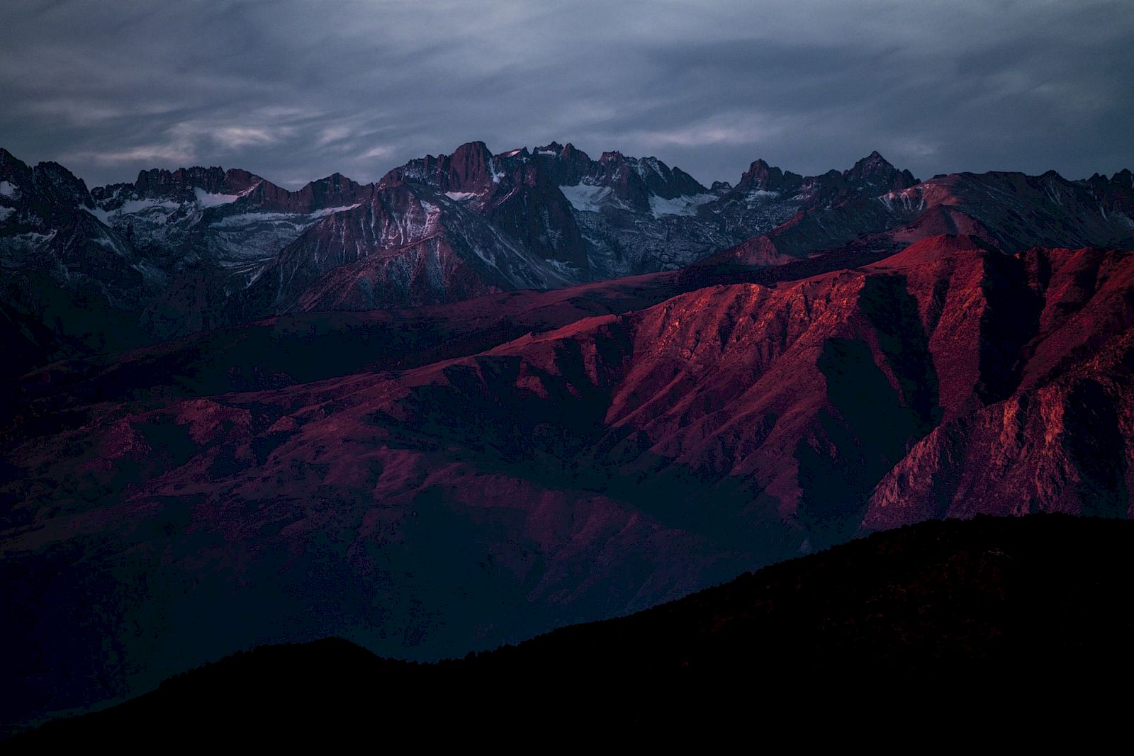 A mountain range with jagged peaks is bathed in a red-purple hue under a dramatic sky.