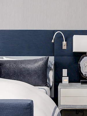 The image shows a modern bedroom with a navy headboard, white bedding, and a nightstand with a lamp and decorative item.