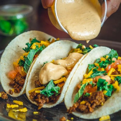 Three soft shell tacos filled with ground meat, lettuce, tomatoes, and cheese are being topped with a creamy sauce on a plate.