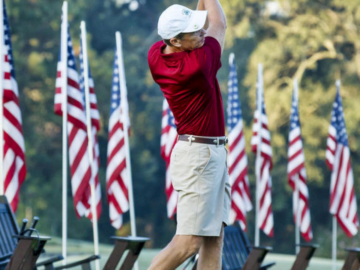 A person in a red shirt and white cap swings a golf club, surrounded by chairs and multiple American flags in the background.