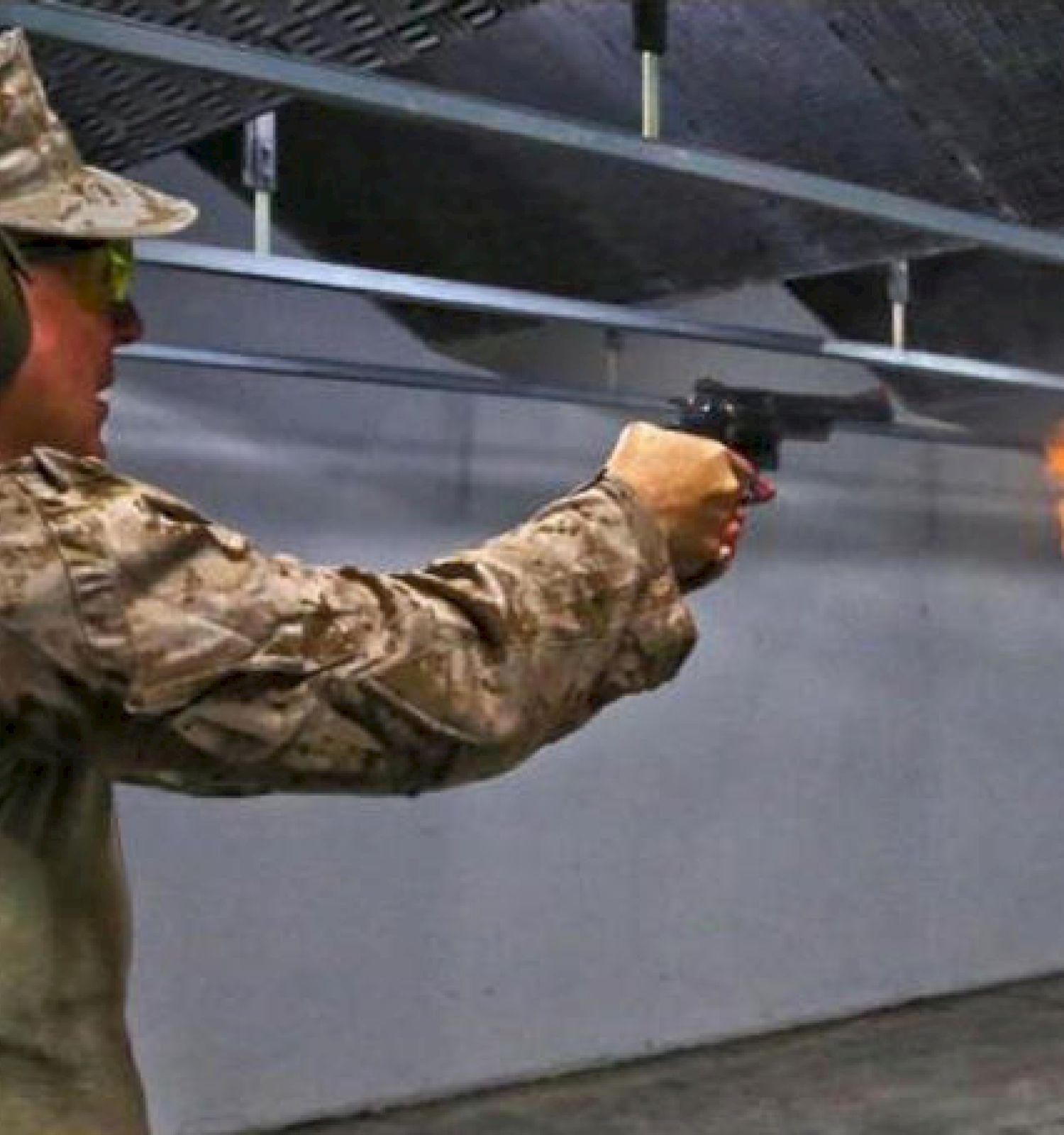 A person in military attire is firing a handgun at an indoor shooting range, with a visible muzzle flash.
