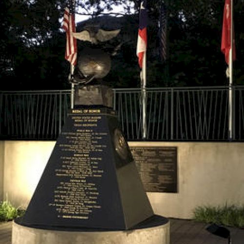 This image shows a monument with engraved text, topped by a helmet and eagle, and is flanked by three flags in a landscaped setting.