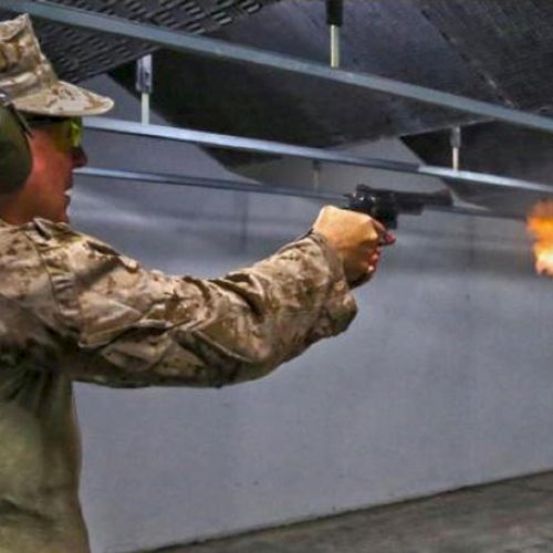 A person in military uniform is firing a handgun indoors, with visible muzzle flash and wearing hearing protection.