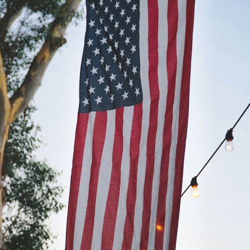 An American flag hangs vertically outdoors, surrounded by trees and string lights, creating a festive atmosphere.