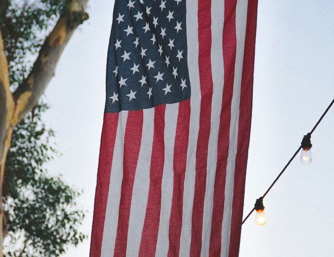 An American flag hangs vertically outdoors, surrounded by trees and string lights, creating a festive atmosphere.