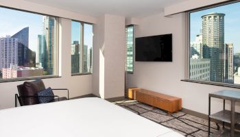 A modern hotel room with large windows offers a city view, featuring a bed, armchair, wall-mounted TV, and minimalistic decor.