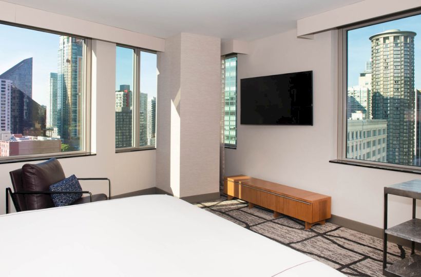 A modern hotel room with large windows offers a city view, featuring a bed, armchair, wall-mounted TV, and minimalistic decor.