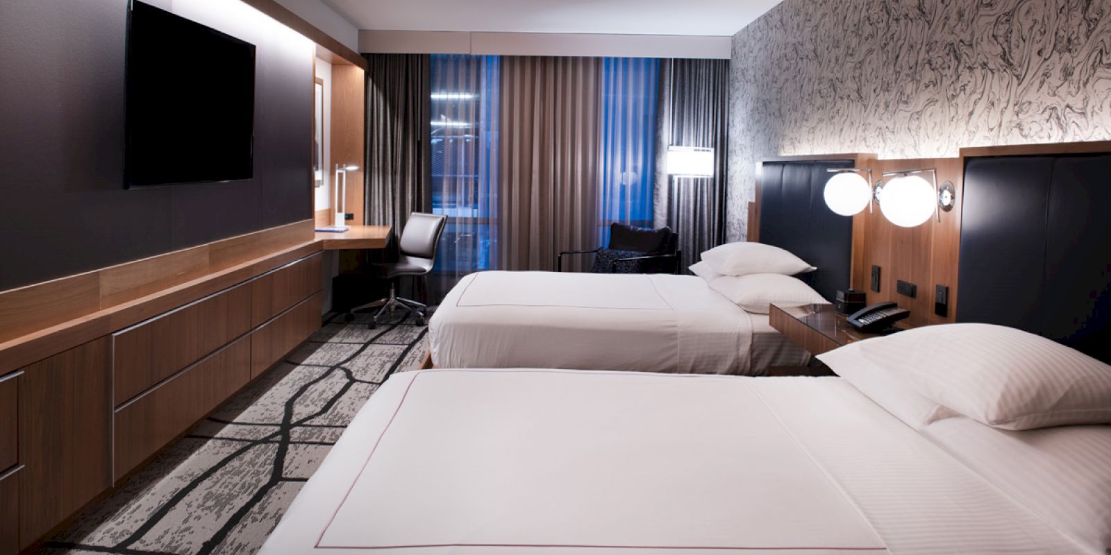 The image shows a modern hotel room with two beds, a wall-mounted TV, a desk, and stylish lighting and decor.
