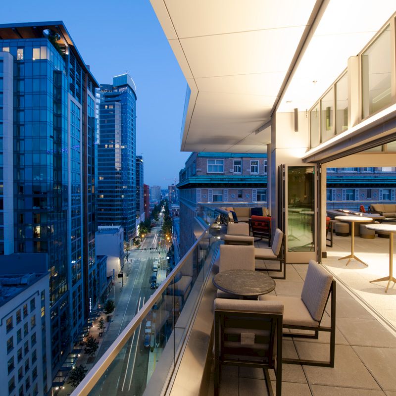 An urban balcony overlooking a city street at dusk, featuring outdoor seating and high-rise buildings with illuminated windows.