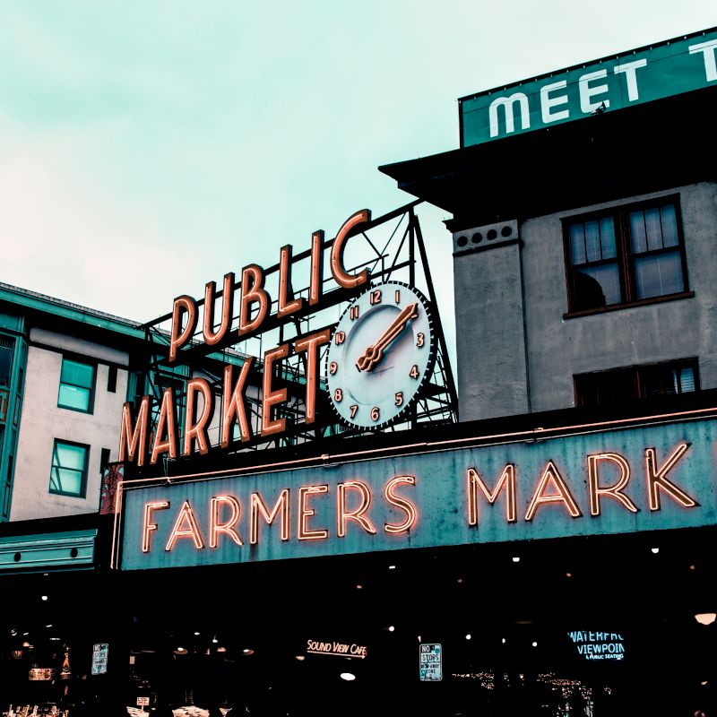 The image shows a neon sign for a Public Market and Farmers Market, with a clock, on buildings under a cloudy sky.