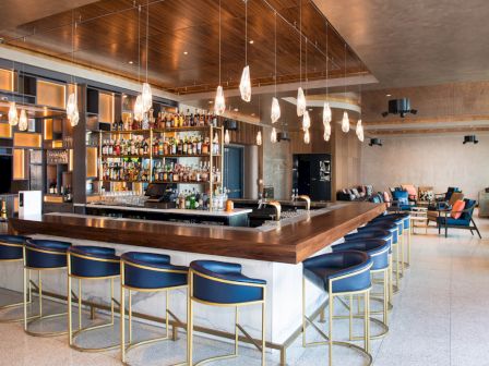 A modern bar with blue stools, a stocked liquor shelf, and hanging lights. There's a seating area in the background.