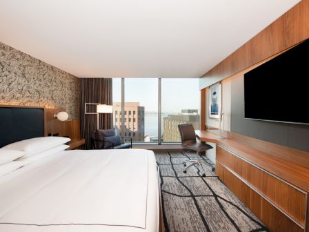 A modern hotel room features a large bed, desk with chair, flat-screen TV, and floor-to-ceiling windows with a city view.