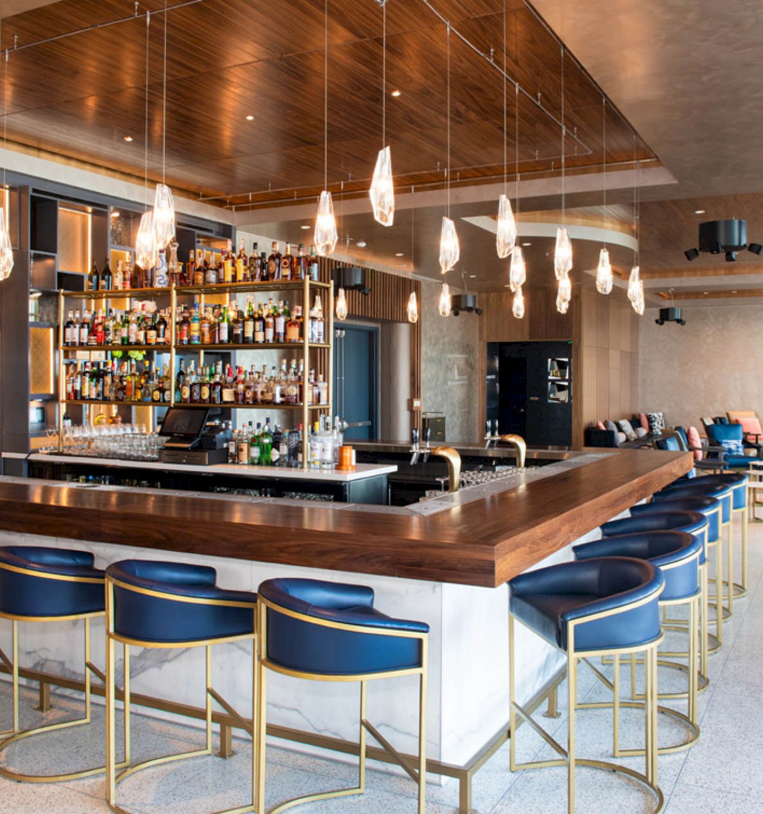 A modern bar with a wooden counter, blue stools, and pendant lights. Shelves stocked with bottles and lounge seating in the background.