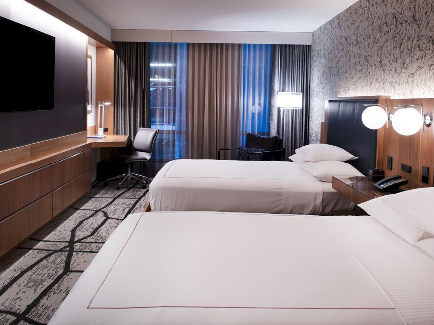 A hotel room with two beds, a TV, a desk, and modern decor, featuring a patterned wall and ample lighting.