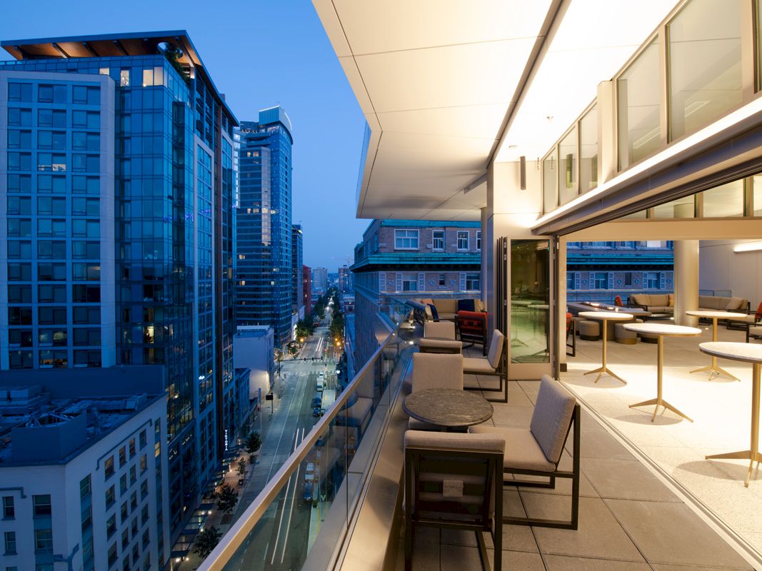 The image shows a modern city view at dusk from a terrace with tables and chairs, overlooking buildings and streets below.