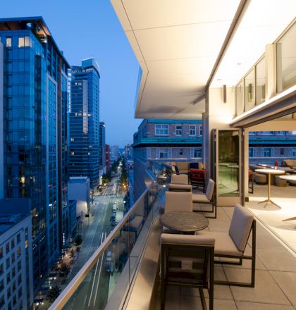 Modern cityscape from a balcony at twilight, featuring tall buildings, street view, and outdoor seating area.