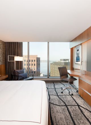 A modern hotel room with a large bed, wall art, a flat-screen TV, a workspace, and a view of city buildings from the window.