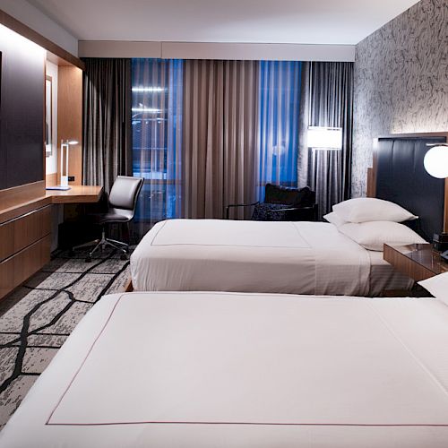 The image shows a modern hotel room with two beds, a desk, a large TV, ambient lighting, and patterned wallpaper.