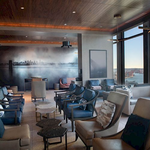 A modern lounge with comfortable seating, large windows offering an ocean view, and a wall mural of a city skyline.