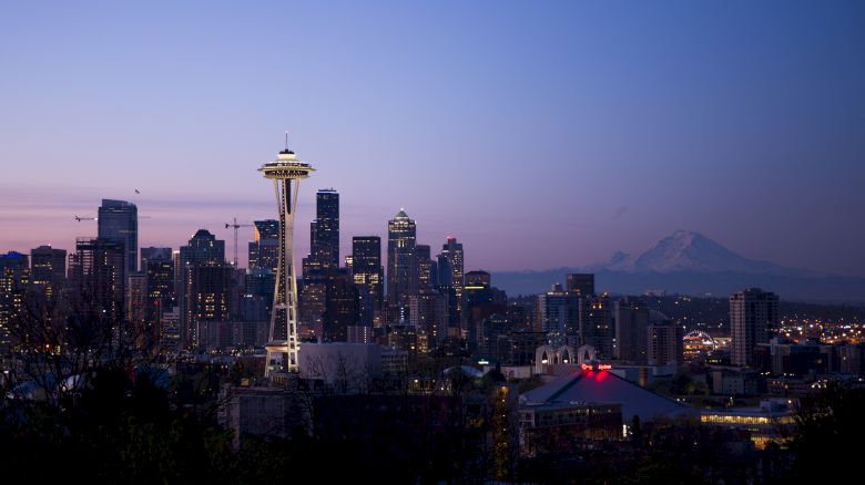The image shows the Seattle skyline at dusk, featuring the Space Needle and Mount Rainier in the background, creating a scenic urban view.