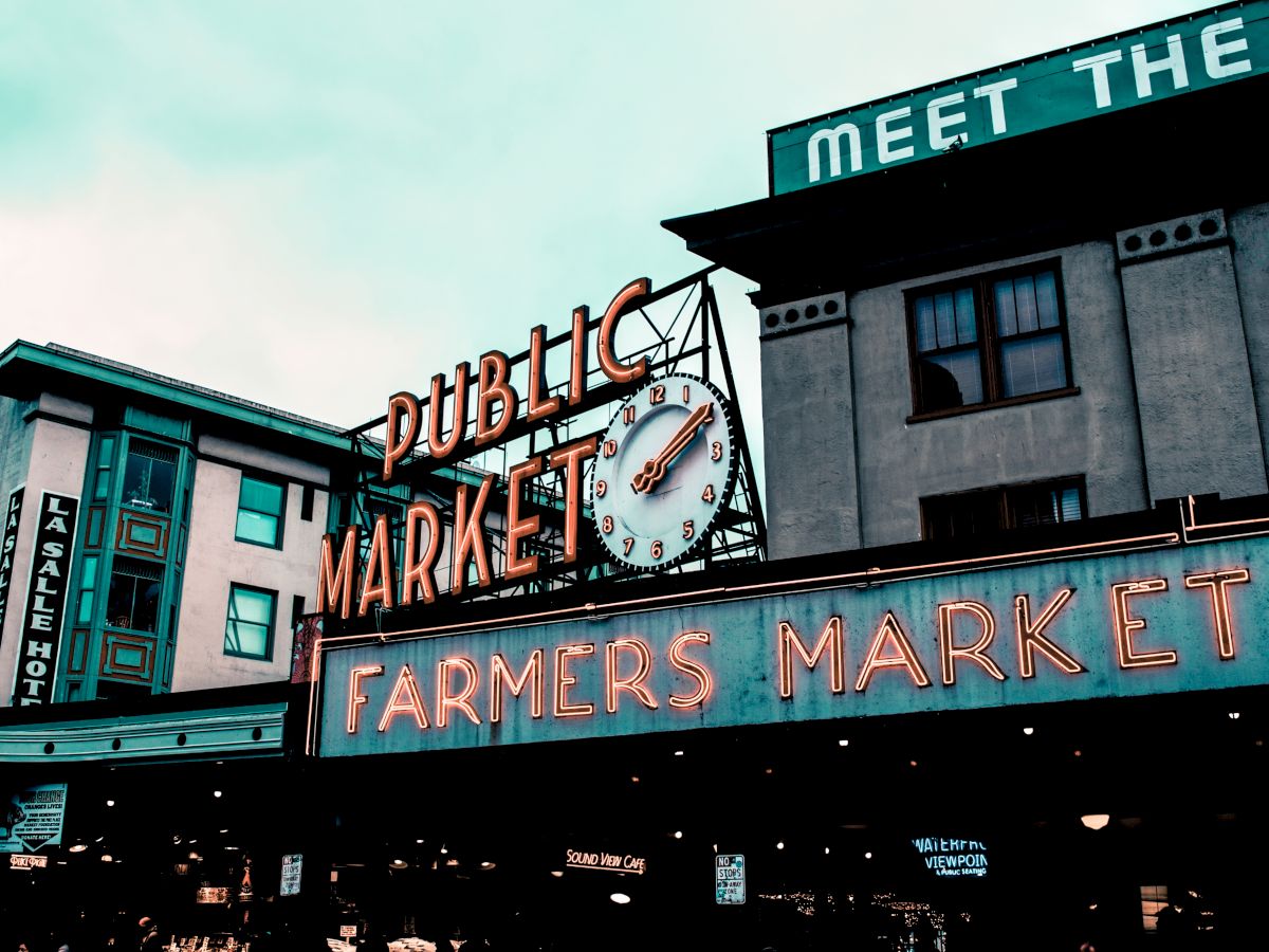 An outdoor market scene with neon signs for "Public Market" and "Farmers Market," featuring a clock and surrounding buildings.
