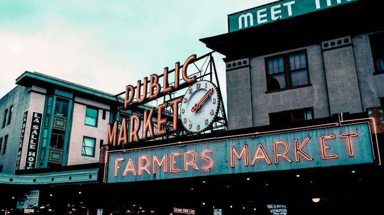 An outdoor market scene with neon signs for "Public Market" and "Farmers Market," featuring a clock and surrounding buildings.