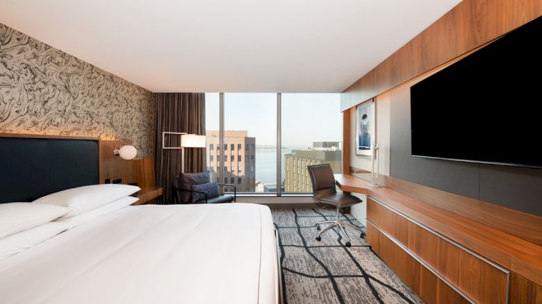 A modern hotel room with a large bed, desk, chair, wall-mounted TV, and a window view of urban buildings and water.
