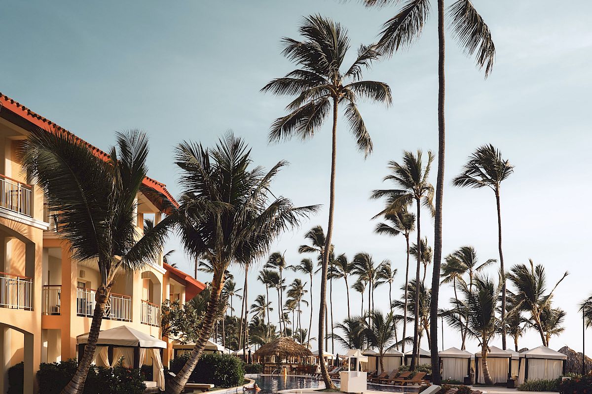 A tropical resort scene with palm trees, a clear pool, and sun loungers arranged near orange-roofed buildings under a blue sky.
