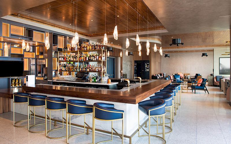 A modern bar with blue stools, a wooden counter, stylish lighting, and shelves stocked with various bottles in a spacious setting.