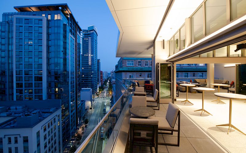 A modern outdoor terrace at dusk overlooking a city street and tall buildings, featuring seating and tables under warm lighting.