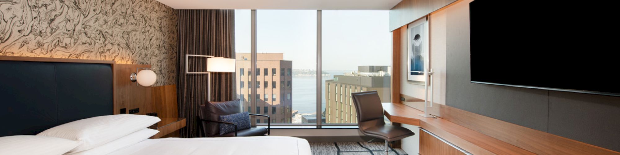 A modern hotel room features a large bed, wall-mounted TV, desk, chair, and cityscape view through wide windows.